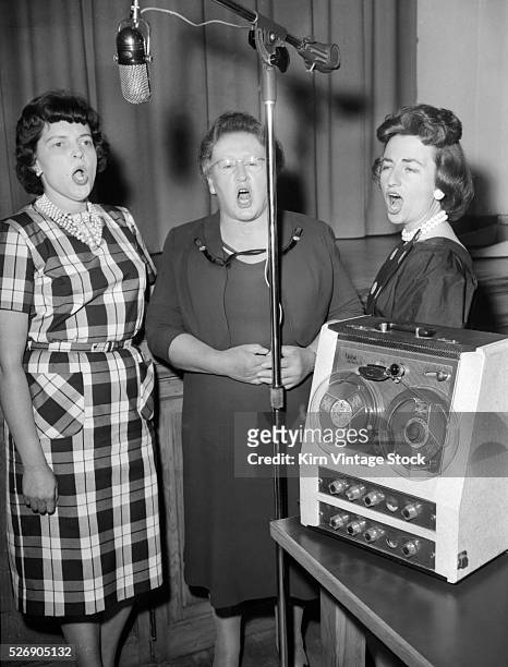 Four women singers belt out a tune in a studio using a reel to reel tape recorder.