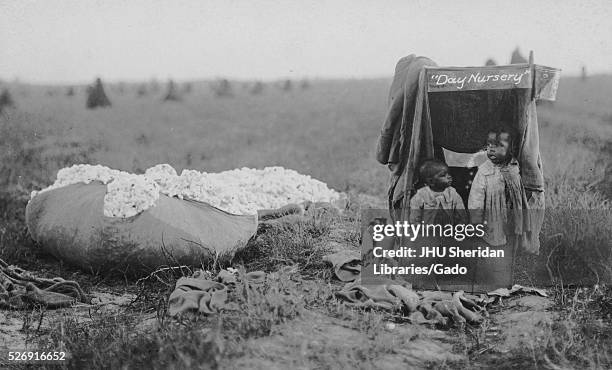 Two young African American children wearing light clothing standing in wooden structure labelled "Day Nursery" with blankets, standing next to big...