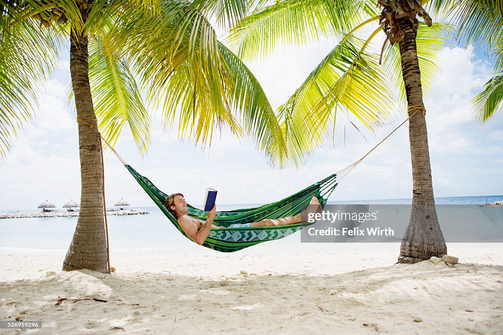 Portrait of man relaxing in hammock on beach reading book