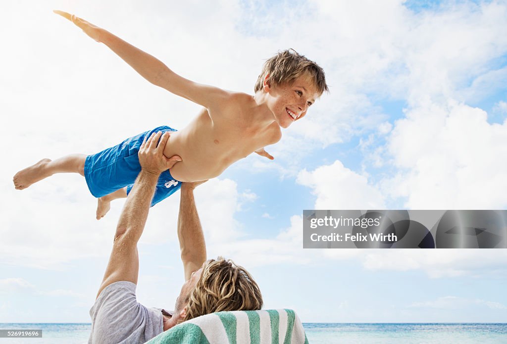 Father holding his son (10-12) mid-air while relaxing on deckchair on beach