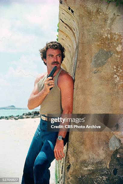 American actor Tom Selleck, in character as private investigator Thomas Sullivan Magnum, hides behind a seawall with an automatic pistol at the ready...