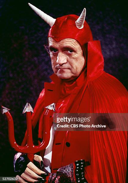 Wry American comedian and actor Bob Newhart looks into the camera with a dour expression as he wears a humorous devil costume replete with red cape,...