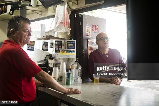 Barman and a customer watch Alejandro Garcia Padilla, governor of Puerto Rico, not pictured, giving a speech on a television screen in a bar in San...