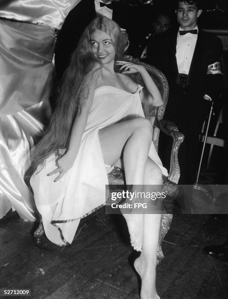 American model Dorian Leigh sits in a Louis XIV chair and wears a diaphanous sheet-like garment and long blonde wig as a young man in a tuxedo with...