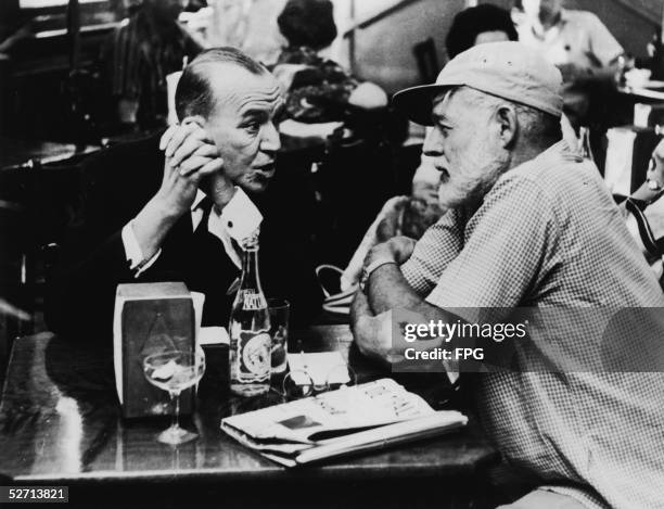 American writer Ernest Hemingway sits with his arms crossed avidly listening to British actor, playwright, and composer Noel Coward at Sloppy Joe's...