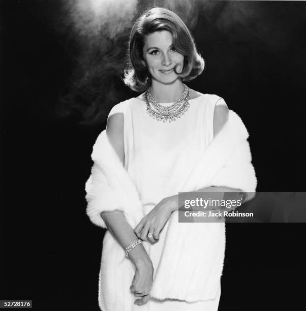 American model and actress Suzy Parker poses with a white fur stole over a white sleeveless dress and wears diamond jewellery, mid 1960s.