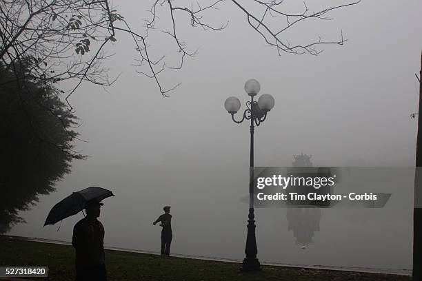 An elderly Vietnamese gentleman stretches on a misty morning beside Hoan Kiem Lake, Hanoi, with the Thap Rue Pagoda visible through the mist. For a...