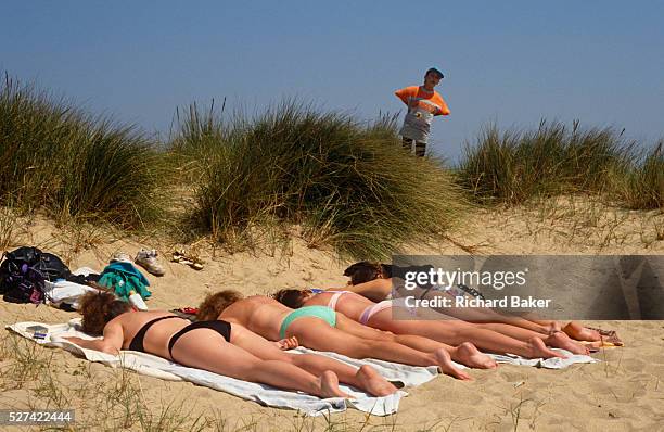 Young boy peers over a clump of vegetation to spy on four beautiful women who are all lying face-down in a sandy dune near the seaside resort of...