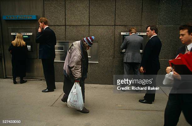 An elderly homeless man walks slowly past a Barclays Bank cash dispenser at which business people are either queueing or typing in their PIN numbers...