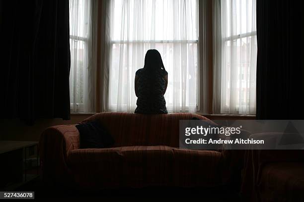 Young Asian woman suffering from domestic violence stands alone in the bay window of her home.
