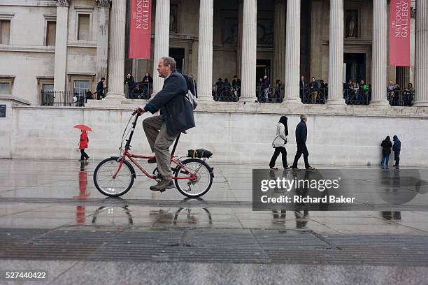 Londoners and cyclist pass-by on the pedestrian pavement in Trafalgar Square in central London. During seasonal rainfall, the pavement stones are...