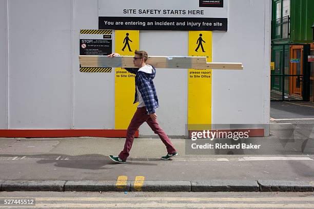 South London youth carries timber over his shoulder past the site entrance of the regeneration project at Elephant & Castle, London borough of...