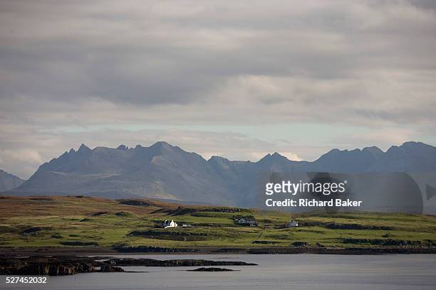 Across the calm waters of a Scottish bay, isolated houses and crofts sit before the dramatic Cuillin Mountains that rise up in the distance on the...