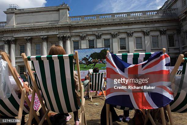 Watching live TV coverage of Equestrian events, British spectators and other sports fans sit in summer deckchairs at the old Royal Naval College,...