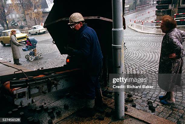 Coal delivery man deposits chunks of brown coal into the cellar via a conveyor belt for an elderly lady who stands outside in the bitter cold wearing...