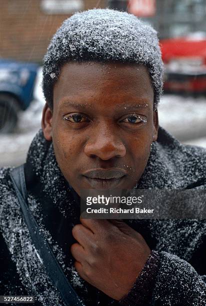 Man of Afro-Caribbean birth, clutches at his scarf to keep out freezing temperatures during a cold snap in south London. Falling snow has settled on...