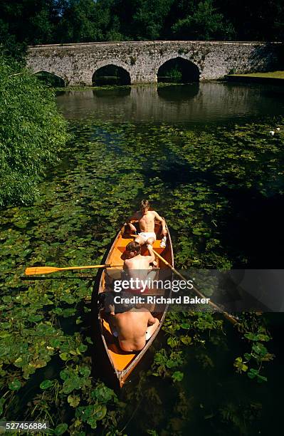 It is mid-day on the narrow stretch of river, green lilly pads float on its surface and in unbder a fierce sun overhead, three young men are lazily...