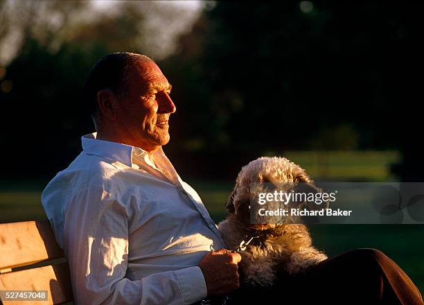 Middle-aged man, possibly in his late-fifties, holds the lead of his pet dog on a park bench in London England. Afternoon sun filters through nearby...