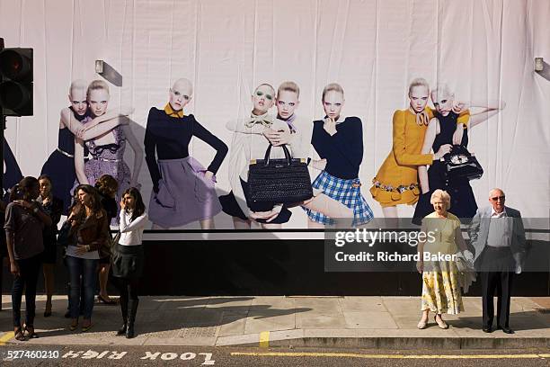 Young women on a shop hoarding with pedestrians and an elderly couple about to cross Brutoin Street in central London. An image of young and older...