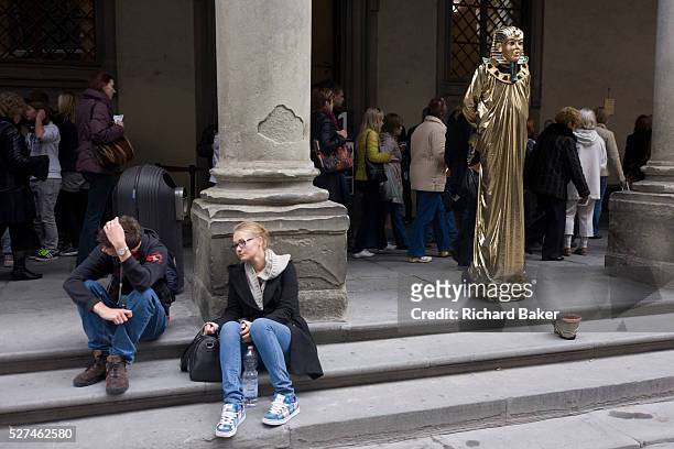 Tired and disappointed tourists and a pretend Egyptian pharaoh busker stand awaiting custom in Florence's Piazza degli Uffizi. In the darker covered...