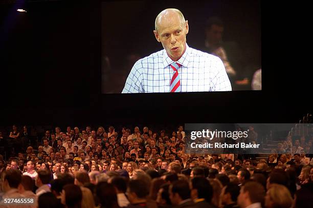 Chairman of Ernst & Young Mark Ottey peers down on his employees on a giant screen, addressing his loyal audience of E & Y staff who have congregated...