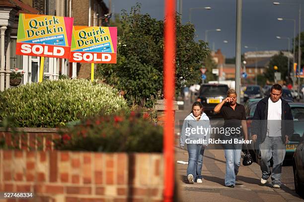 Young family walk gloomily past property Sold signs in a street at Grays, Essex England. Passing the prominent signs that bear the name of Quirk...