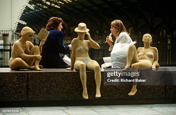 Female colleagues enjoy a chat over an alfresco lunch in the city alongside an art installation of women at the beach. Rather suggestively we see,...