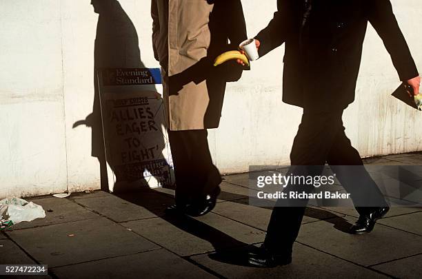 Businessman strides along a London street holding a banana whose shadow appears to be part of another man's anatomy. As strong sunlight shines on...