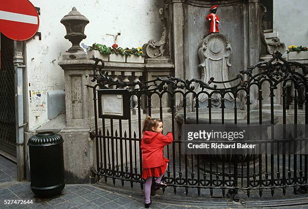 Young girl holds railings at the site of Brussels' famous landmark, the Mannekin Pis statuette, dressed in red. A red theme appears from the...