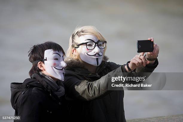 Couple wearing Anonymous masks pose for their own selfie photo. Holding up a smartphone to take the picture, the person on the right touches the...