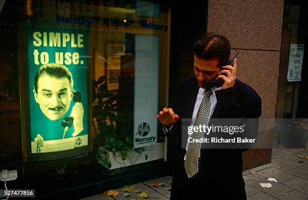 In front of an ad for Mercury, the 90s mobile phone network provider, a city worker uses his mobile phone in a London street. Actor Harry Enfield was...