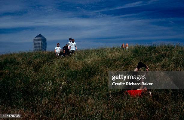 Near children playing with a goat and a man laying with just his knees showing, a woman exercises her hamstrings in long grass on an embankment near...