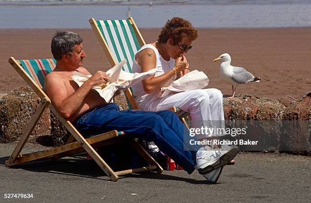 Holiday couple sit in deck-chairs to enjoy their chips wrapped in paper, the traditional way for eating fish and chips while at the seaside. The...