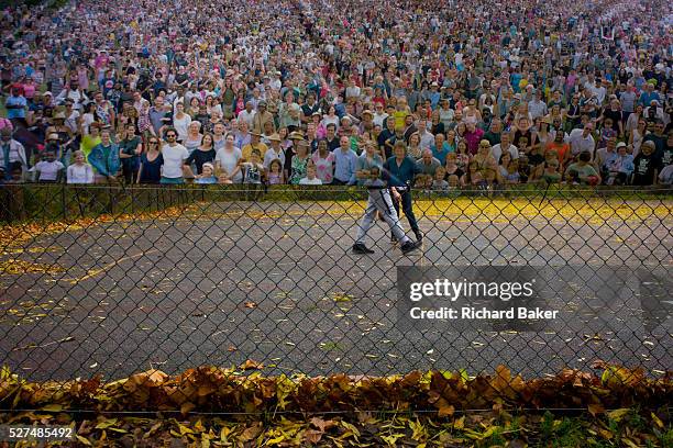 The lower legs of two boys playing basketball, partially intersected by a poster of a crowd - in a south London park. We look through the wire fence...