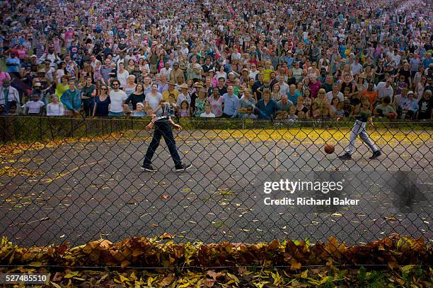 The lower legs of two boys playing basketball, partially intersected by a poster of a crowd - in a south London park. We look through the wire fence...