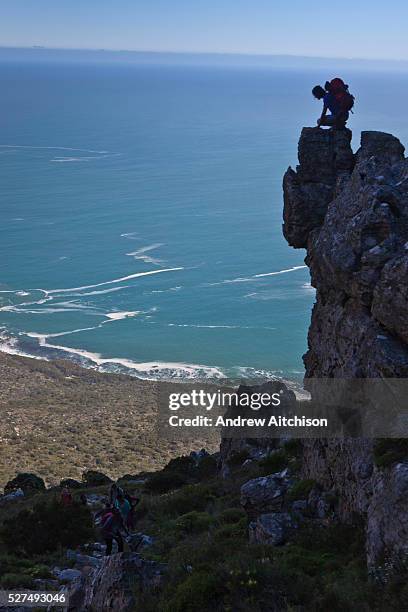 Hike instructor leans over a mountain cliff edge to talk and encourage along the children on the expedition on Table able Mountain, Cape Town, South...