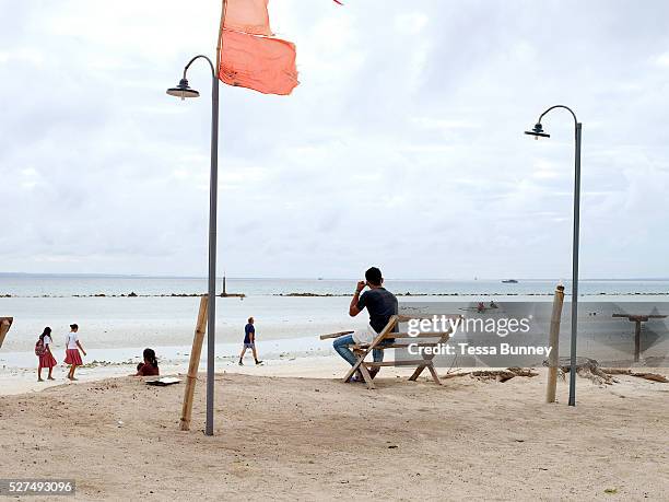 Beach scene in Santa Fe, Bantayan Island, The Philippines. A man sitting on a wooden bench watches while school girls walk along the beach and...