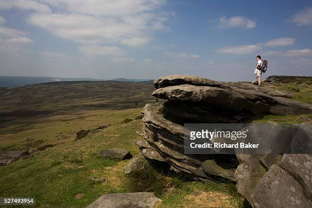 Lone female walker rests on top of Stanage Edge gritstone cliffs. From the summit of this escarpment, we see a wide landscape of Gritstone geology on...