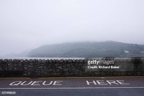 Queue Here sign on the road at the Corran Ferry crossing on Inverscaddle Bay, Ardgour, Scotland. On a bleak and grey summer evening, with low clouds...