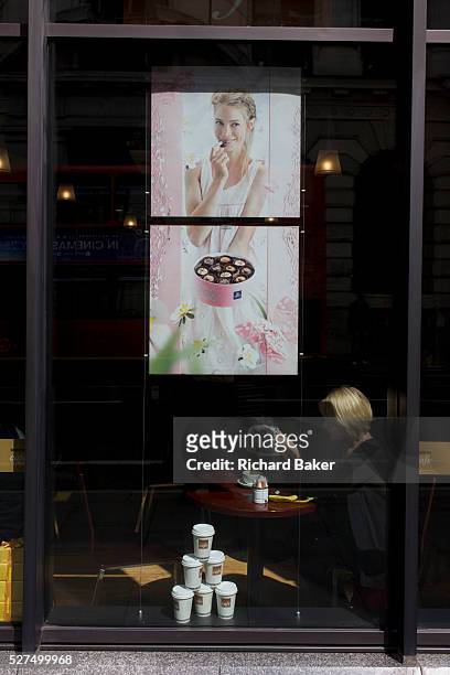 Businesswoman sits in sunlit window beneath poster showing quality Belgian chocolates, in Manon Cafe on King William Street in the City of London,...