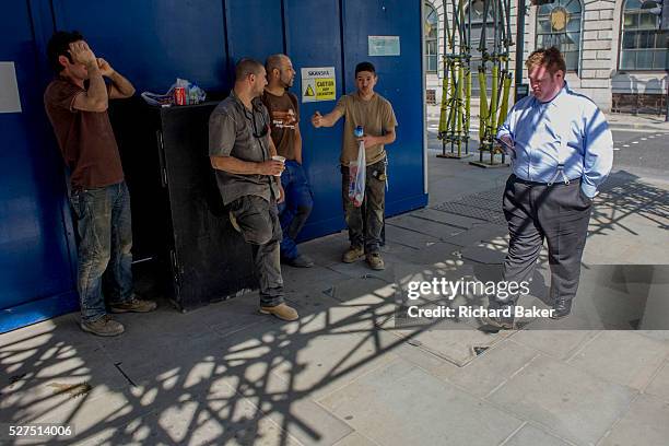 In the shadow of scaffolding, a large office worker in braces uses his smartphone as he passes a group of resting construction workers during a hot...