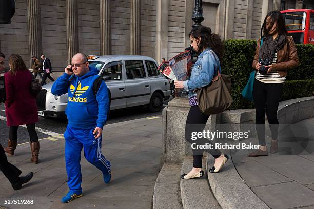 Man wearing a bright Adidas tracksuit-type of sports clothing walks and talks passing the pillars of the Bank of England. On an otherwise dull street...