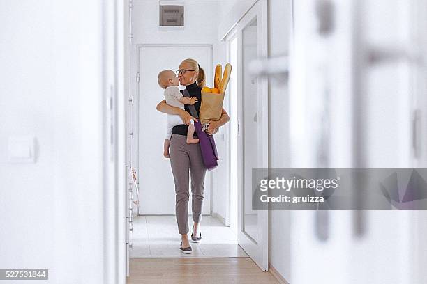 young mother enters home, carrying her baby and grocery bag - family hugging bright stockfoto's en -beelden