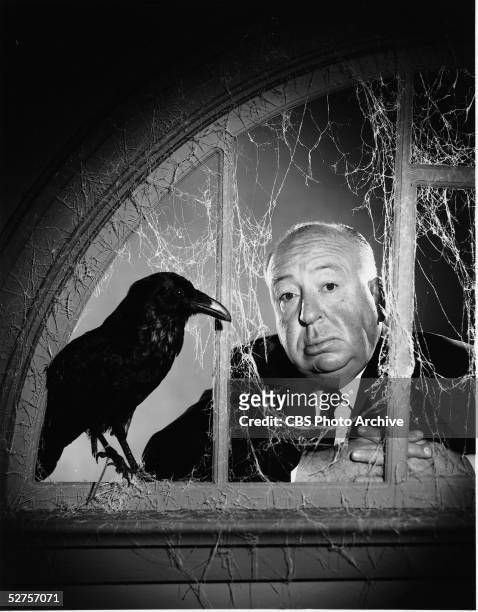 Portrait of English-born American film director Alfred Hitchcock as he stands behind a fake window which is covered with fake cobwebs and on the sill...