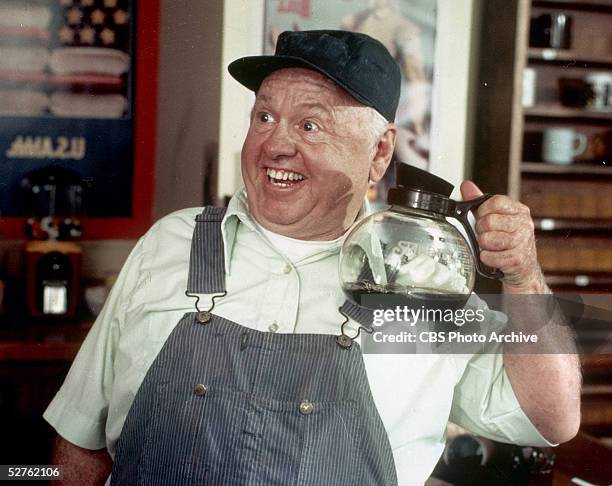 American actor Mickey Rooney, dressed in striped overalls and a cap, holds up a pot of coffee in a scene from the television movie 'Bill,' directed...