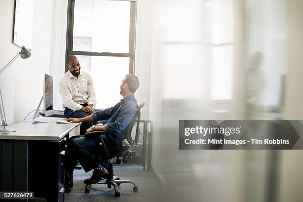 office life. a man leaning back in an office chair talking to a colleague sitting on the edge of the desk. - 2 people back asian stock-fotos und bilder