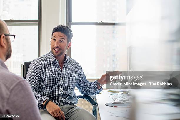office life. two men seated talking and one using his hand to gesture towards a computer monitor. - open collar stock pictures, royalty-free photos & images