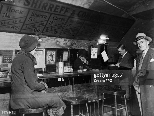 Woman sits on a barstool as several men stand in a winebar, Great Britain, 1930s. Above the bar is a board which displays the prices of available...