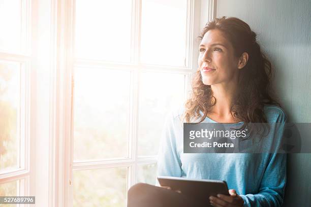 pensierosa giovane donna che tiene compressa digitale dalla finestra - tre quarti foto e immagini stock