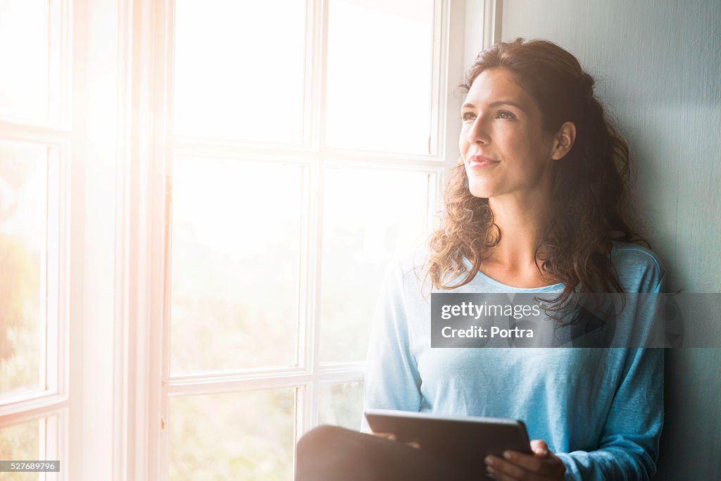 Thoughtful young woman holding digital tablet by window